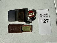 Top view of the Zippo lighter, Ronson lighter, Boy Scouts pin, Saks cardholder and card along with maxsold lot number ticket