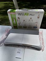 Photo of the Wii Fit Plus balance board in front of its original box showing branding and fitness poses.