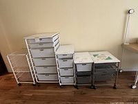 Front view of five distinct Container Store Elfa rolling storage racks with varying drawer configurations in white and gray color on wood floor against beige wall.