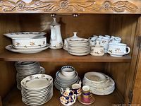 Photo showing set of plates, cups, serving dishes, cream and sugar set with floral patterns in a wooden cabinet.