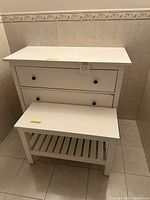 White Ikea dresser with two drawers and black knobs next to small white table in tiled room