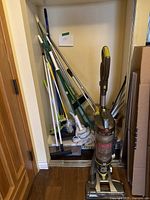 Full view of vacuum and multiple cleaning tools stored upright in a closet space showing mops, brooms, sweepers, and the vacuum.