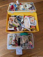 Top-down view of yellow and clear plastic containers filled with assorted sewing supplies including thread spools, needles, buttons, tape measures, and pins.