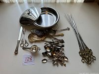 Assorted silver plated bowls, spoons, serving utensils, and skewers arranged on a table showing quantity and condition.