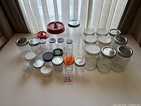 Lot of 26 assorted glass canning jars with various lids displayed on table in front of window.