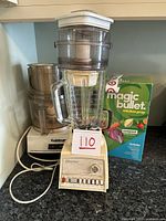 Vintage Osterizer blender with glass jar, Cuisinart food processor, and boxed Magic Bullet appliance on a kitchen counter.