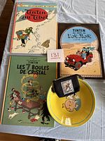Photo of three Tintin hardcover books: Tintin au Tibet, Les 7 Boules de Cristal, Tintin au Pays de L'Or Noir, yellow bowl with Tintin character inside, black small pouch with Tintin label