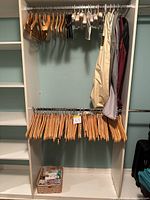 Photo of two rows of wooden hangers and satin padded hangers on rods inside a white closet unit. Several garment bags hanging on the side. Basket with moth protection cakes and balls on the bottom shelf.