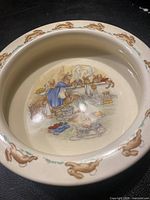Top-down photo showing inside of beige ceramic bowl decorated with Bunnykins rabbit illustrations depicting cooking scene, surrounded by rabbits along rim.