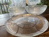 Three clear glass dishes with textured floral patterns: two bowls of different sizes and one large platter, shown on wooden surface with natural light.