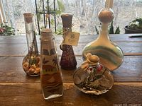 Five glass decorative bottles with colorful sand art and paraffin oil candle placed on wooden table with window background