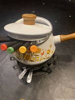 Full view of floral fondue pot with lid and 6 multi-colored forks inserted, showing floral pattern and wooden handles.