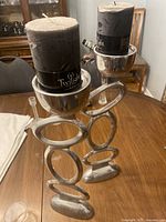 Two dark gray pillar candles with black Twilight Collection Rustic bands, set in shiny silver metal holders with interconnected oval shapes as bases, placed on a wooden table in a dining room setting.