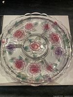 Top-down photo of clear glass serving tray with raised floral designs in pink, purple, and green on surface. Tray has scalloped edges and divided serving sections.