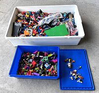 Large white storage bin and blue bins filled with assorted Lego bricks, baseplates, and minifigures including bags of parts
