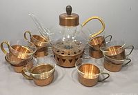 Front view of the glass teapot with woven wicker handle, copper lid, infuser, copper warming stand beneath, surrounded by 10 glass cups with copper holders, wicker and metal handles visible.
