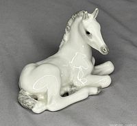 Glossy white porcelain foal figurine in lying position, front-left angle view.
