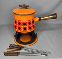 Bright orange fondue pot with floral decoration, wooden handle and lid knob, displaying eight fondue forks with wooden handles lying in front, set on matching orange base with missing candle holders.
