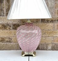 Front angled view of pink swirl Murano glass lamp with brass base and white floral patterned shade wrapped in plastic.