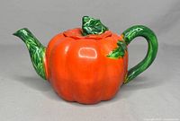 Side view of vintage tomato-shaped teapot showing green leaf handle and spout, orange body with lid on top.