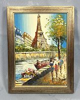 Framed oil painting showing Eiffel Tower and Paris riverside with people, trees, and buildings under bright blue sky.