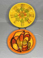 Photo of two Poole Pottery Delphis plates, one yellow with floral design, one orange with abstract design, standing upright.