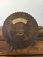 Front view of the antique brass circular calendar showing the engraved floral pattern, years, and dates.