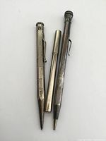 Three antique silver mechanical pencils laid side-by-side on white background. Left pencil has hexagonal barrel with textured patterns and clip. Center pencil is smooth and cylindrical with separate cap. Right pencil is slim and tapered with clip. All exhibit tarnishing and surface wear. No packaging visible.
