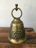 Front view of large vintage brass bell showing circular handle, floral relief, and Latin text.