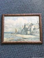 Full view of framed watercolour painting depicting a coastal scene with stone buildings, cliffs, and a boat on the shore.