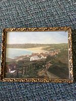 Framed watercolour painting showing a coastal village with houses, bay, and hills.