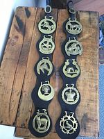 Full view of two black leather straps laid on a wooden surface, each with five brass plaques depicting horses and various symbols.