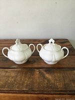 Pair of white porcelain lidded sugar bowls on wooden surface showing front and sides.