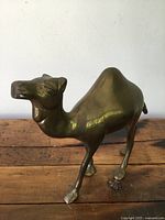 Side and front perspective of vintage brass camel figurine on wooden surface against white wall, showing shape and surface wear.
