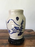 Full view of off-white vintage pottery vase hand painted with blue landscape, displayed on dark carved wooden Asian style stand, showing general shape and decoration.