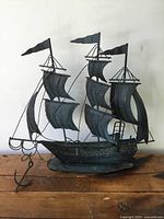 Front view of the vintage metal ship decor showing three masts with flags and curved sail design, resting on oval base with anchor extending from the bow.