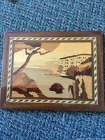 Full view of wooden inlay wall picture showing coastal landscape within a darker wooden frame, set against a blue carpet background.