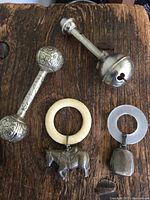 Four antique baby rattles on a wooden surface showing two metal rattles and two ring-shaped rattles with pendants.