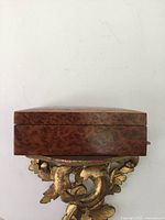 Closed rectangular burl wood dresser box placed on a decorative carved gilded support, showing rich wood grain pattern.