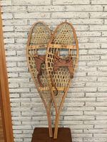 Front view of the pair of antique Canadian snowshoes leaned against a white brick wall, showing the wooden frames, rawhide webbing, and leather bindings.