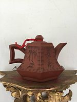 Front side of the reddish-brown hexagonal Yixing clay teapot showing Chinese calligraphy and geometric handle with red cord on lid.