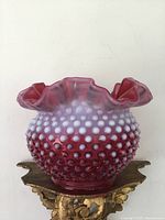 Front view of cranberry hobnail vase on decorative stand showing hobnail texture and ruffled rim.