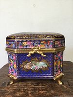 Front view of the porcelain lidded box showing detailed hand painted floral decorations, central classical figure scene, gold gilding, and ornate gold feet.