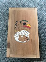 Top view of rectangular cedar wood box lid showing hand-painted North West Coast Indigenous artwork of a bird with a yellow beak above a small sloth wrapped in a white shell.