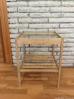 Front view of a small side table with a wooden frame and a glass top, set against a white brick wall. The bottom shelf consists of several wooden slats evenly spaced.