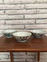 Front view of three antique Chinese hand painted bowls arranged side by side on a wooden table with a white brick wall background.