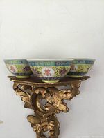Photo showing three painted Chinese tea bowls with red character marks on bases, placed on ornate gold stand.