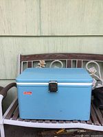 Blue vintage Coleman cooler on metal bench against light green siding. Cooler is rectangular with blue finish, white lid trim, metal latch front, and side handles. Visible wear including scratches and scuffs.