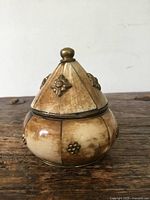 Front view of the antique inset panel lidded box showing conical lid with brass floral decorations and rounded base with matching brass floral details.