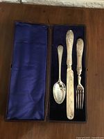 Top view of vintage silver plated knife, fork, and two spoons arranged in original leather fitted box with blue velvet and purple satin lining.